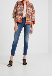 Paksu oranssi neulecardigan, jossa on geometrisia kuvioita, yhdistettynä istuviin sinisiin farkkuihin ja mustiin fringe-helmalla varustettuihin avointen varpaiden kenkiin.