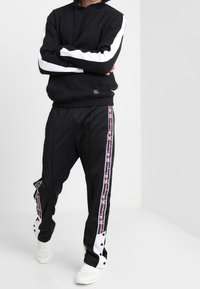 Svart hoodie med vita ärmar, matchad med svarta träningsbyxor med vita och röda sidostreck och tryckknappar. Vita sneakers.