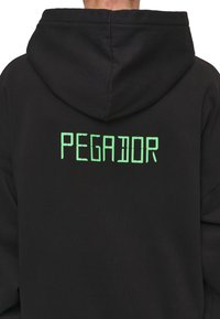 Sweat à capuche noir avec une capuche, arborant un texte réfléchissant vert "PEGADOR" au dos dans une police rectangulaire de style numérique. Tissu doux et texturé.