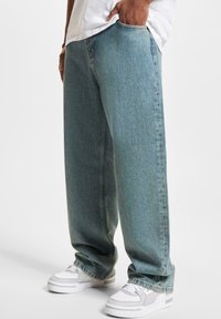 Jeans de mezclilla azul claro con un corte relajado, piernas rectas y costuras visibles. Combinados con zapatillas deportivas blancas y grises.