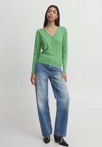 Grüner Pullover mit langen Ärmeln, V-Ausschnitt und Knopfakzenten, kombiniert mit hellblauen weiten Jeans und schwarzen Absatzschuhen.