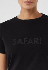 Czarna koszulka w dopasowanym kroju, z napisem "SAFARI" w błyszczącej wersji. Materiał wydaje się miękki i gładki, z klasycznym okrągłym dekoltem.