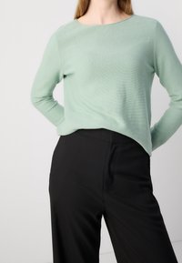 Mintgrüner Pullover mit Rundhalsausschnitt und langen Ärmeln, aus einem weichen Stoff gefertigt. Kombiniert mit maßgeschneiderten schwarzen Hosen mit Taschen.