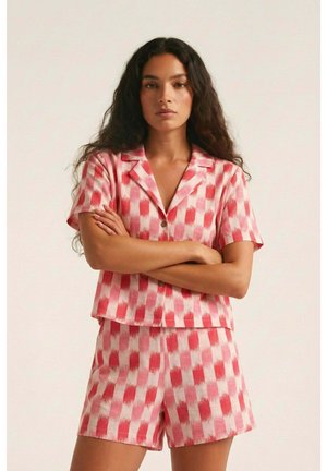 Mujer con el cabello largo y oscuro que lleva una camisa de manga corta a cuadros a juego en rojo y rosa, y pantalones cortos, de pie con los brazos cruzados.