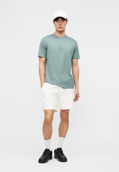 Νέος άνδρας που φοράει λευκό καπέλο, πράσινο μπλουζάκι με λογότυπο, λευκό σορτς, λευκές κάλτσες και μαύρα αθλητικά παπούτσια, στέκεται με το ένα χέρι στην τσέπη.