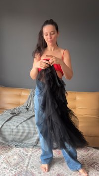 Tessuto di tulle nero con volants tenuto in mano, abbinato a un top rosso e jeans blu. Sofà in pelle beige e tappeto a fantasia sullo sfondo.