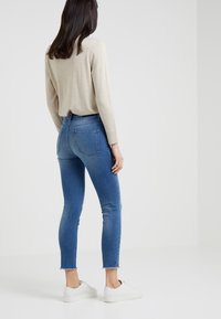 Smala jeans i medelblå denim med en fransad nederkant, kombinerat med en ljusbeige långärmad topp och vita sneakers. Bakifrån.