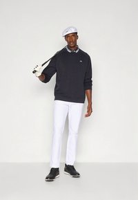 Στολή γκολφ με σκούπερ σκούτερ, λευκό παντελόνι και μαύρα παπούτσια. Το μοντέλο κρατά ένα γκολφ ρόπαλο, φορώντας λευκό καπέλο και γάντι.