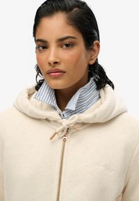 Haut à zip beige clair avec cordon de serrage, col rayé pastel bleu et blanc contrastant, et logo "Superdry" en relief sur la poitrine.