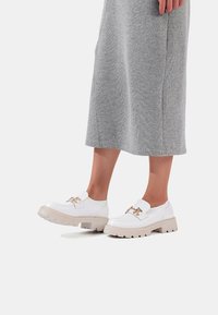 Scarpe platform bianche con finitura in pelle liscia, dotate di una fibbia dorata e una robusta suola in gomma beige. Vestito grigio testurizzato visibile.