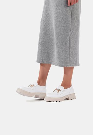 Scarpe platform bianche con finitura in pelle liscia, dotate di una fibbia dorata e una robusta suola in gomma beige. Vestito grigio testurizzato visibile.