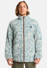 Chaqueta de forro polar en azul claro con un patrón abstracto blanco, cuello alto y cremallera marrón. Presenta un parche con el logo en el pecho.