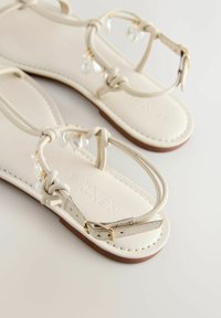 Bézs lapos szandál vékony pántokkal, bokapántos csat, és díszítő gyöngyszerű gyöngyökkel a fonott elülső pánton, fehér felületen.
