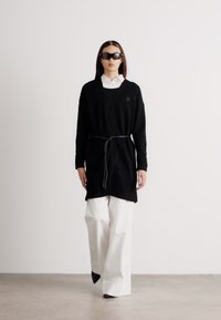 Μαύρο πουλόβερ με μακριά μανίκια και κορδόνι στη μέση, φορεμένο πάνω από λευκό πουκάμισο με γιακά, συνδυασμένο με φαρδιά λευκά παντελόνια.