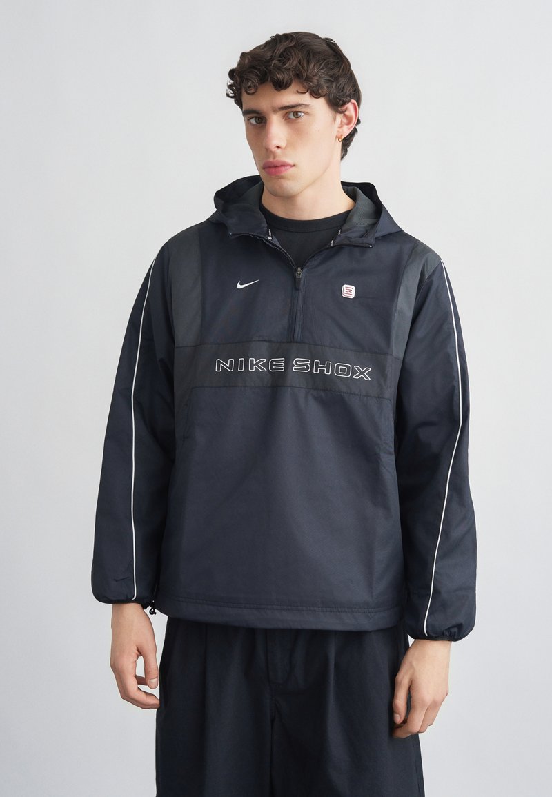 Νεαρός με σγουρά σκουρόχρωμα μαλλιά, φορέας ενός μαύρου αδιάβροχου Nike Shox με λευκή ρίγα, στέκεται μπροστά σε ένα απλό φωτεινό φόντο.