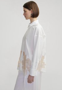 Camisa blanca de algodón con bordado floral dorado a lo largo del lado inferior, con collar clásico y mangas largas.