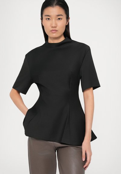 Vrouw met steil zwart haar, gekleed in een nauwsluitend zwart T-shirt met korte mouwen en een bruine broek, staat met één hand in haar zak.