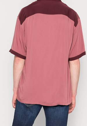 Achteraanzicht van een persoon die een losvallend, kortärmelig poloshirt in stoffig roze draagt met een donker wijnrode inzet bij de schouders en mouwboorden, gecombineerd met donkerblauwe jeans.