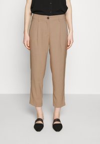 Pantalones cortos beige con pliegues frontales, combinados con zapatos negros de punta afilada y una blusa negra sin mangas metida por dentro.