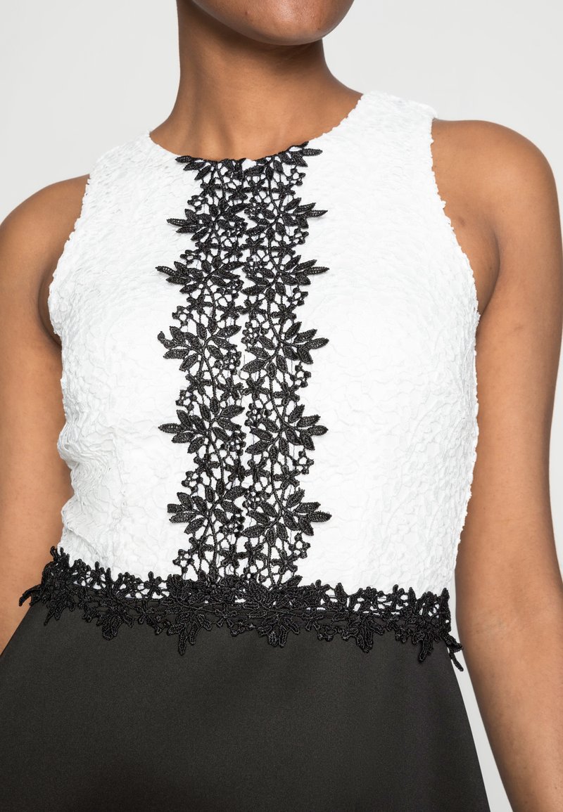 Schwarz-weißes Kleid mit einem strukturierten weißen Oberteil, das in der Mitte mit einem schwarzen Spitzenmuster verziert ist, und einem schlichten schwarzen Rock, der an der Taille verziert ist.