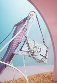 Sininen käsilaukku, jossa valkoinen "Barbie"-logokuvio, kolmionmuotoinen, kiiltävä hopeinen ketjuhihna ja näyttävä pyöreä logokoriste.