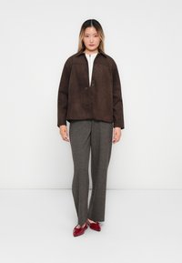 Brun semsket skinnjakke med krage og frontlukking med knapper, parret med grå teksturerte bukser og røde slip-on sko. Enkel design, nøytrale farger.