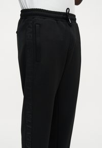 Μαύρο παντελόνι φόρμας με κορδόνι στη μέση, πλαϊνές τσέπες και διακριτική κάθετη επωνυμία στο αριστερό πόδι, φορεμένο με λευκό πουκάμισο.