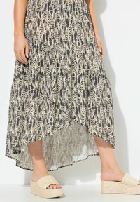 Maxikleid mit einem gestuften Design, das ein beiges und schwarzes abstraktes Muster aufweist. Kombiniert mit hellen, strukturierten Keilsandaletten.