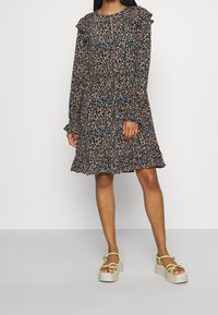 Femme portant une robe florale à manches longues multicolore avec des volants, associée à des sandales compensées jaune pâle sur un fond uni.