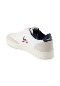 Witte lage sneaker met beige suède accenten, rode hanenlogo aan de zijkant, marineblauwe binnenvoering en 'le coq sportif' tekst op de hieltab.