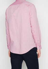 Langärmliges, rosa Hemd mit Knöpfen und einem subtilen Muster, mit abgerundetem Saum und Knopfmanschetten. Der Stoff wirkt leicht und strukturiert.