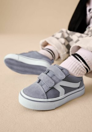 Copil purtând adidași gri cu talpă albă și două benzi velcro, asortați cu șosete albe cu dungi negre și pantaloni cu carouri.