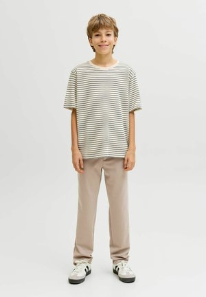 Nasmiješeni dječak stoji okrenut prema naprijed noseći bež i crnu prugastu majicu kratkih rukava, bež hlače i bijele tenisice s crnim prugama.