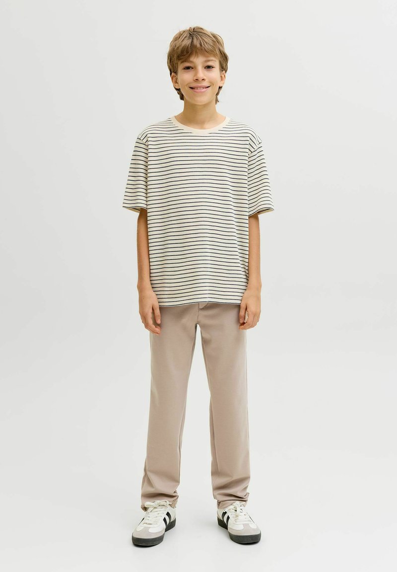 Garçon souriant, debout de face, portant un t-shirt à manches courtes beige avec des rayures noires, un pantalon beige et des baskets blanches à rayures noires.