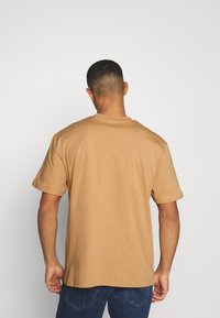 Camiseta de manga corta y cuello redondo en marrón claro, fabricada en suave algodón. Presenta un corte relajado con una textura lisa y un detalle de costura mínimo.