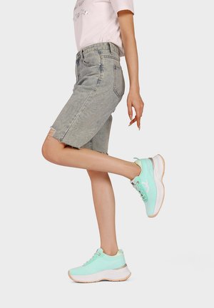 Persona che indossa sneaker verde chiaro e pantaloncini grigi di jeans fino al ginocchio, sollevando una gamba dietro a sfondo uniforme.