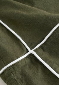 Gros plan sur un tissu vert olive avec un détail de garniture blanche formant une croix à l'intersection des coutures.