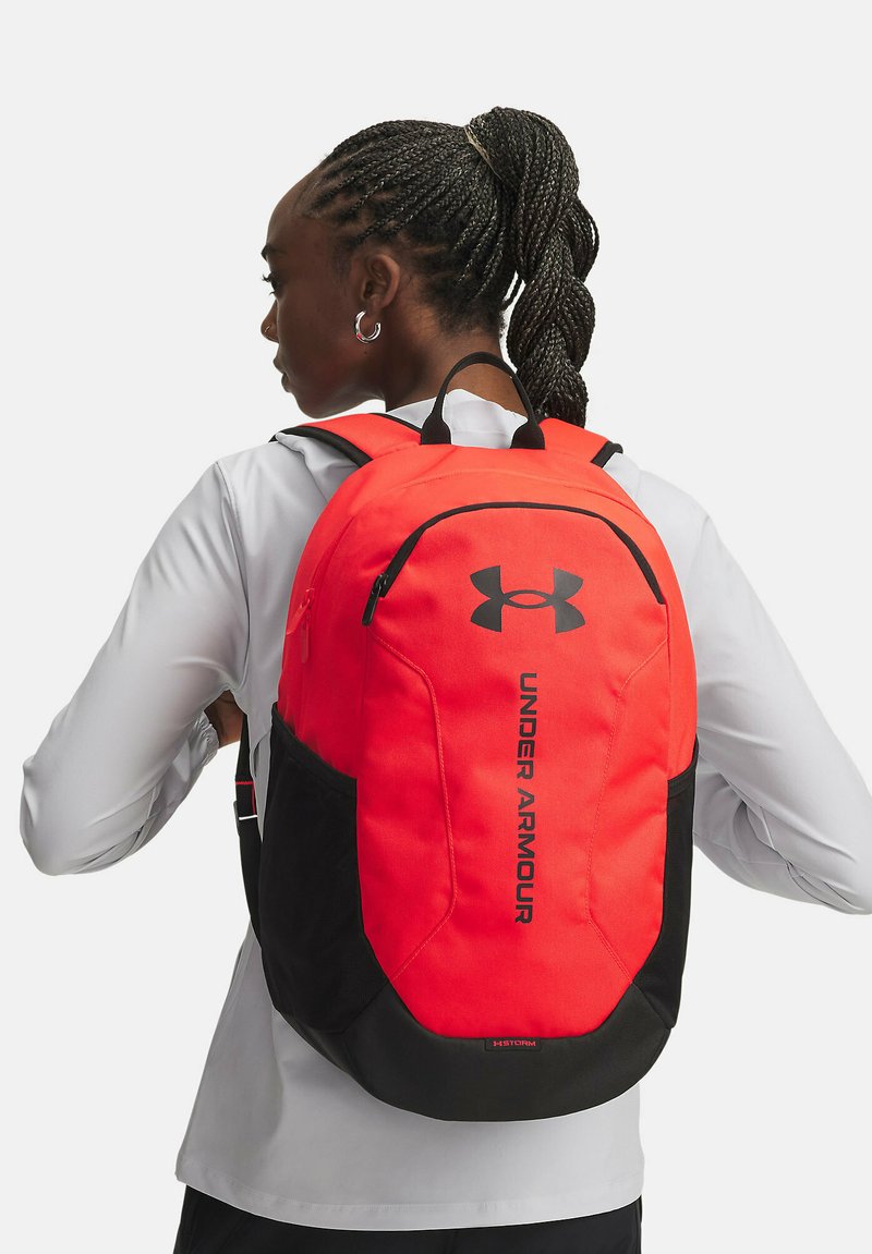 Mochila de Under Armour en rojo y negro con un diseño aerodinámico, correas acolchadas y un logotipo prominente en la parte frontal. Material duradero.