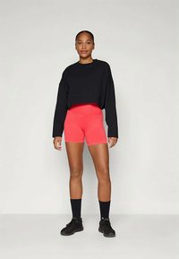 Melnā kroptā džemperis, spilgti rozā šorti, sporta apavi un melnas zeķes. Modelis stāv taisni ar rokām uz gurniem, neitrālā fonā.