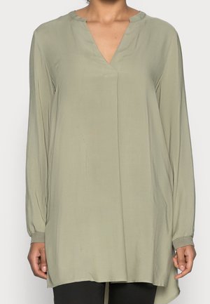 Femme portant un haut tunique ample à manches longues de couleur vert olive, avec un col en V et un devant sans boutons.