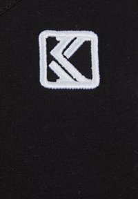 Svart stoff med en glatt tekstur som har en hvit brodert logo innenfor en rektangulær ramme. Logoen inkluderer stiliserte bokstaver "K" og "S."