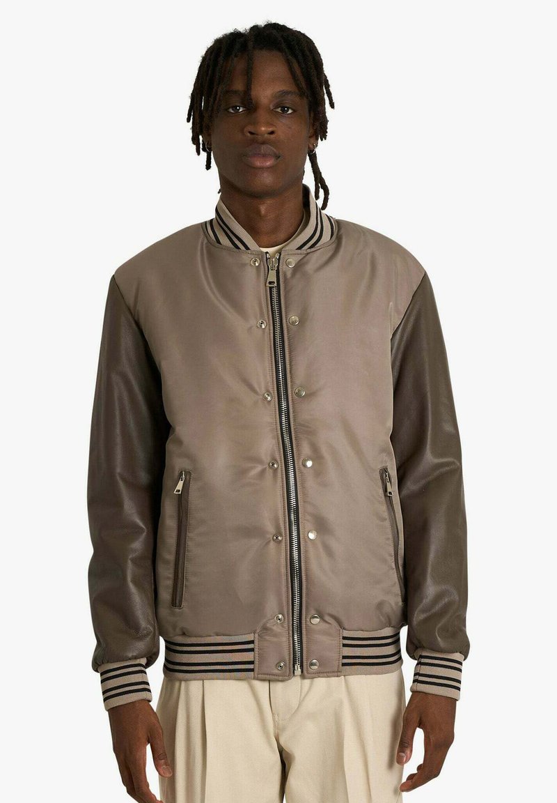 Giacca bomber marrone con tessuto liscio, collo e polsini a costine a righe, tasche con cerniera e chiusura con bottoni a pressione.