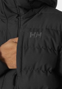 Chaqueta acolchada negra con textura acolchada, que presenta un bolsillo con cremallera en la parte delantera y un logo bordado. Diseño de capucha curvada y elegante.