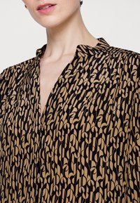 Woman wearing a black and tan patterned blouse with a V-neckline and small standing collar, showing part of her neck and lips.