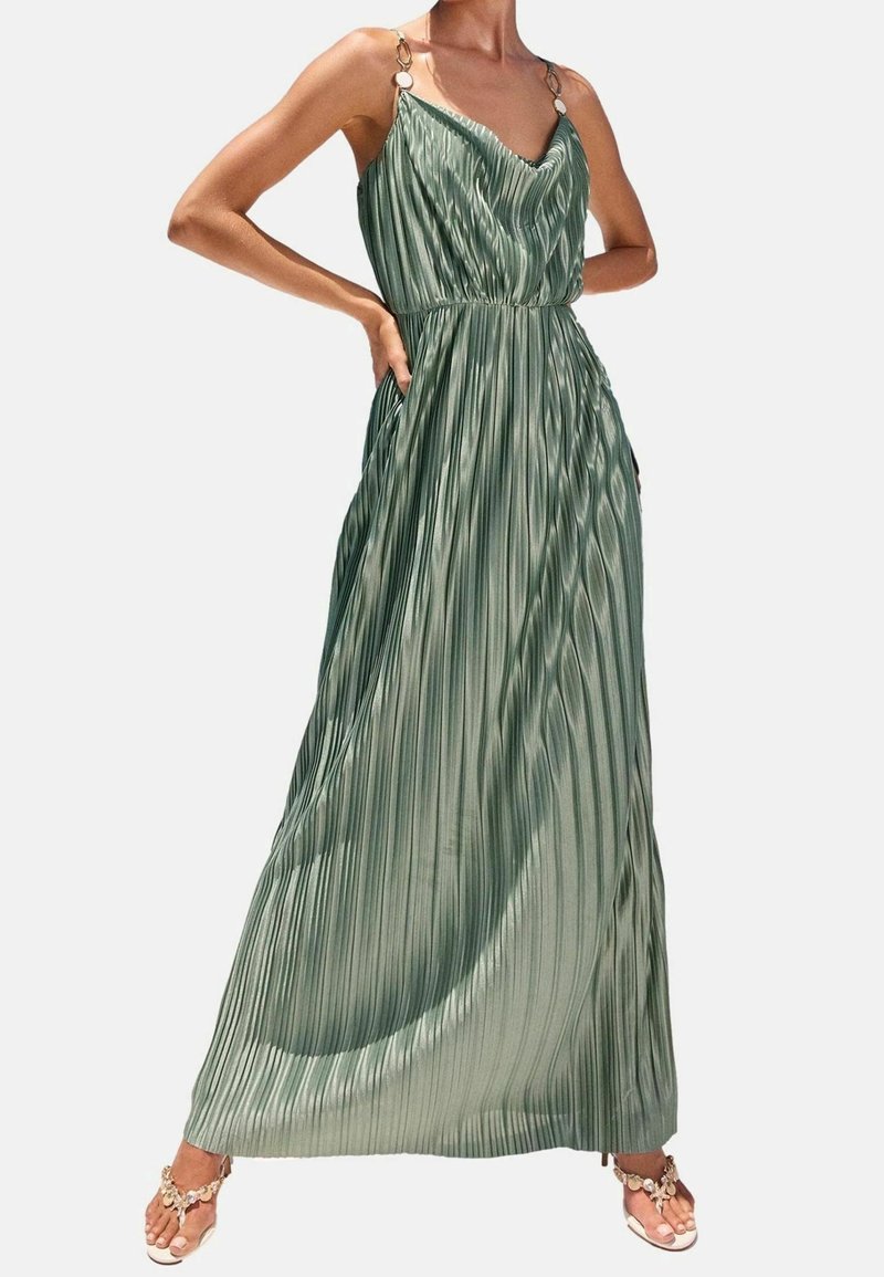 Femme portant une robe maxi plissée métallisée vert sauge avec des bretelles en chaîne dorée et des sandales à bout ouvert ornées de coquillages.