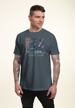 Mand med tatoveringer, iført en mørkeblå T-shirt med Star Wars-figurer og teksten "Kom til den mørke side" i en stiliseret skrifttype.