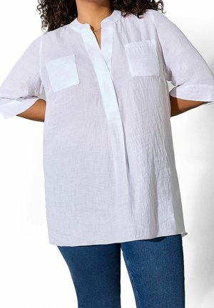 Witte losse blouse met twee voorzakken en driekwart mouwen, gedragen met blauwe jeans, handen achter de rug rustend.