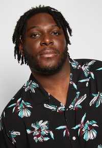 Short-sleeve black shirt featuring large white and turquoise floral patterns. Soft fabric with a collar and front button detail.