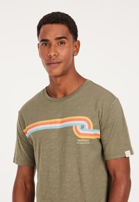 Camiseta de algodón verde de manga corta con un diseño de rayas curvadas multicolor en rojo, naranja y azul, con detalles de logo y etiqueta.