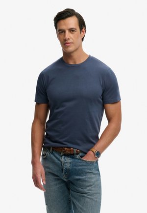 Uomo che indossa una maglietta aderente blu scuro, jeans azzurri, cintura marrone intrecciata e un orologio da polso, in piedi con una mano in tasca.